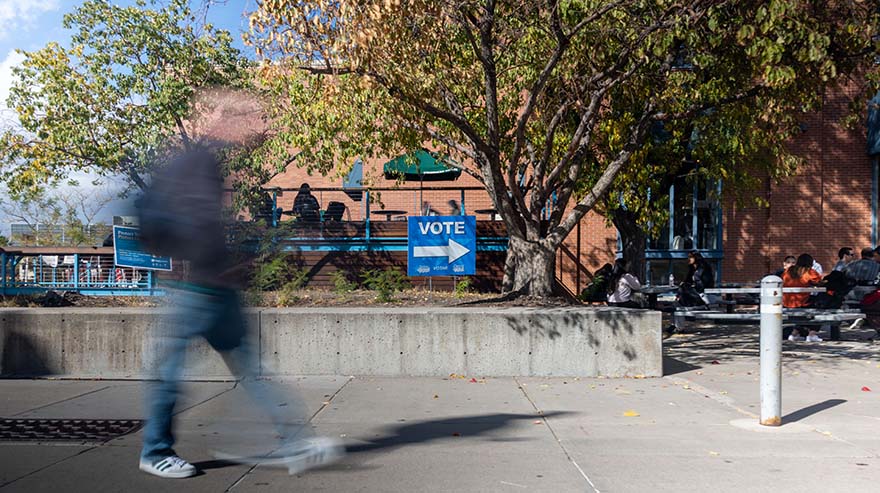 A blurred figure walks past a 'Vote' sign on a college campus with trees and students sitting nearby