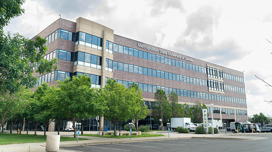 The exterior of the Metropolitan State University of Denver Administration Building, a multi-story brick and glass structure surrounded by green trees and a nearby parking area.