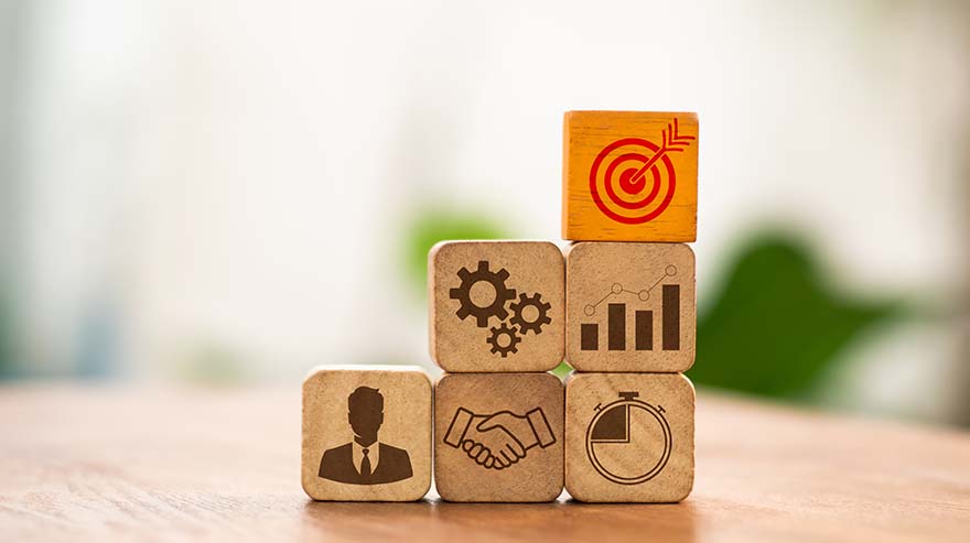 Wooden blocks with icons representing business elements such as a target, gears, a bar chart, a handshake, a timer, and a person in a suit, arranged in a pyramid shape