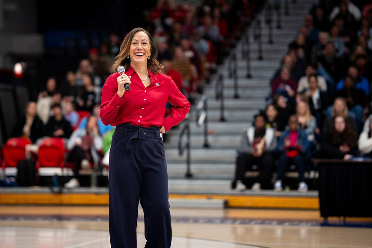 Dr. Davidson in a red shirt holding a microphone on a stage, smiling as she addresses an audience seated in a gymnasium.