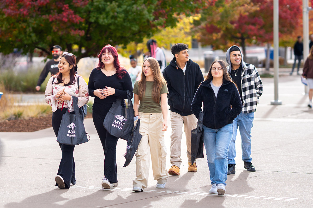 Group of six students walking together outside on a campus, some holding MSU Denver bags.