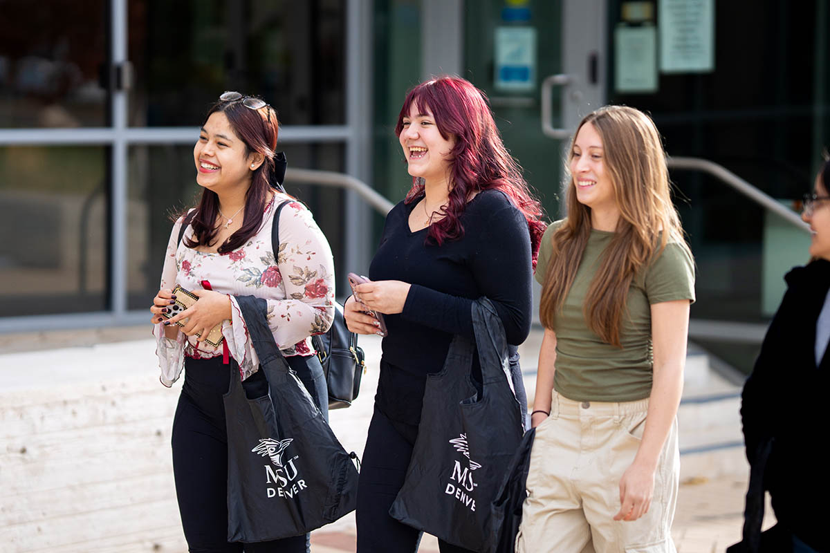 Three students walking and smiling outside on campus with MSU Denver bags.