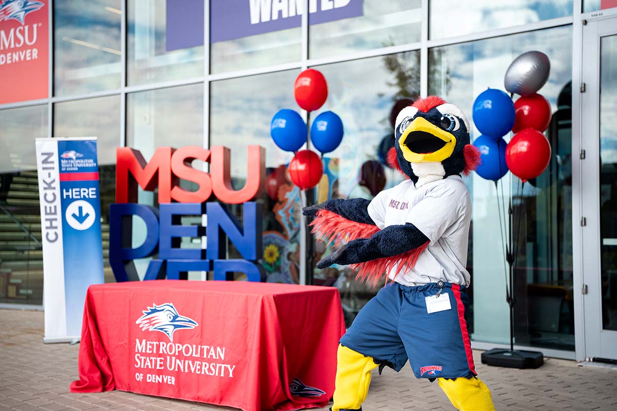 MSU Denver's mascot stands next to a check-in table outside a campus building with decorations.