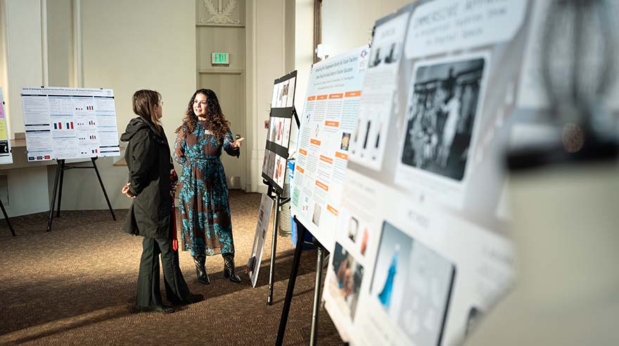 Two people stand in a well-lit room, discussing research posters displayed on easels. The person on the right, wearing a patterned outfit, gestures toward the poster, while the other, in a dark coat, listens attentively. The posters contain various research presentations, including charts, images, and text, set up for a symposium or academic event. The room features soft lighting, large windows, and an elegant atmosphere.