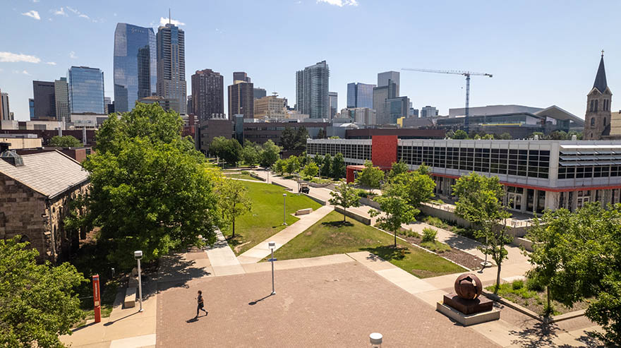 Aerial view of the Auraria Campus in Denver, featuring a brick plaza, modern buildings with a red accent wall, a historic stone structure, green spaces with pathways, and the downtown Denver skyline in the background.