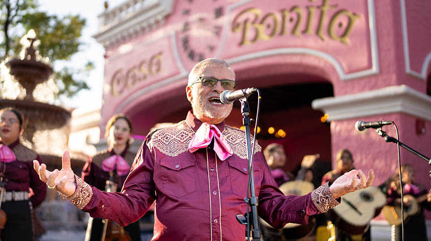 Lorenzo Trujillo, in a maroon mariachi outfit, speaks into a microphone with Casa Bonita's pink building in the background.