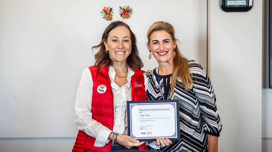 Two individuals smile while holding a certificate for the Roadrunner Shoutout Award. One person wears a red vest with a "Rock the Vote" button and a festive headband, while the other wears a black and white patterned blouse.