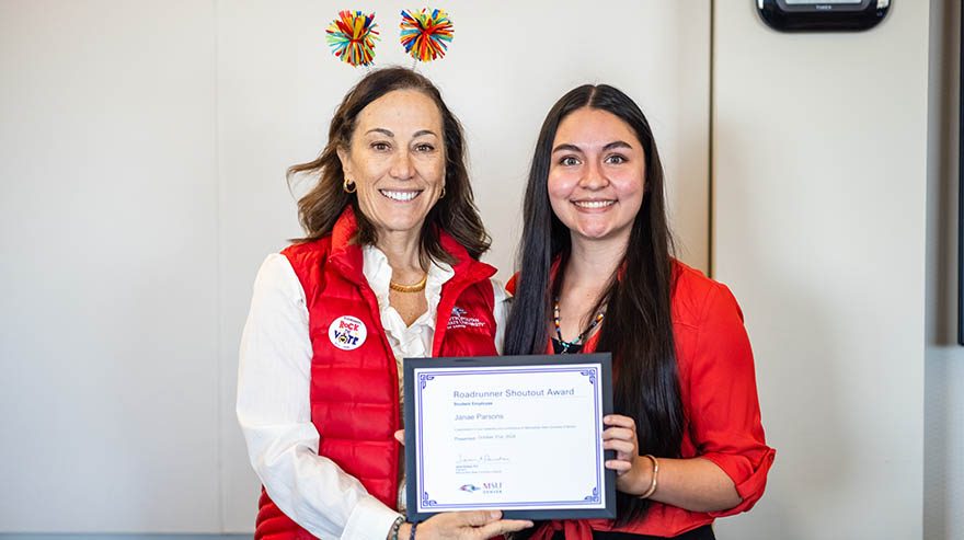 MSU Denver President Davidson  stands with student Janae Parsons  holding a Roadrunner Shoutout Award certificate. Davidson wears a red vest with a "Rock the Vote" button and a festive headband, while the other wears a red blouse.