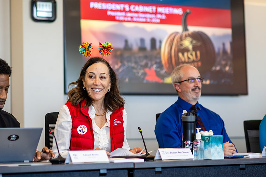 MSU Denver President dons festive headwear during a meeting of her cabinet. She sits in front of an image of a Halloween pumpkin displayed on a TV screen.