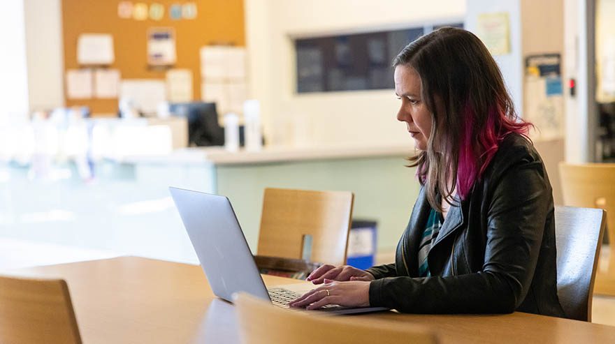 A person with shoulder-length hair dyed with pink streaks, wearing a black leather jacket, sits at a wooden table using a laptop. The background shows a blurred office or academic setting with bulletin boards and office equipment.