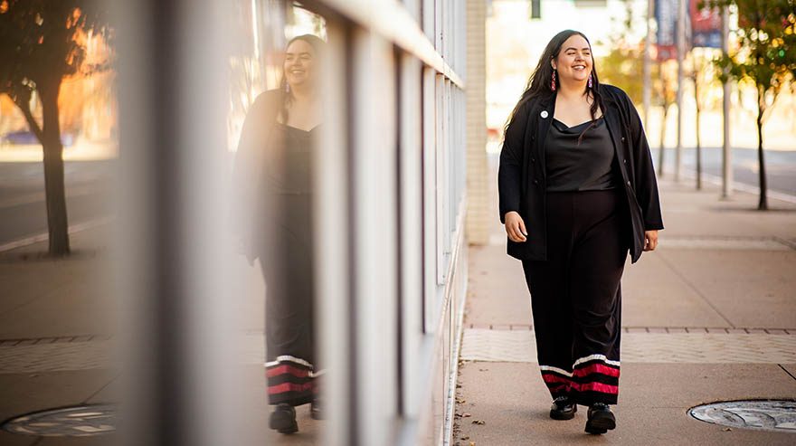 Shayla Bischoff walking on a sidewalk, smiling, with her reflection in a window beside her.