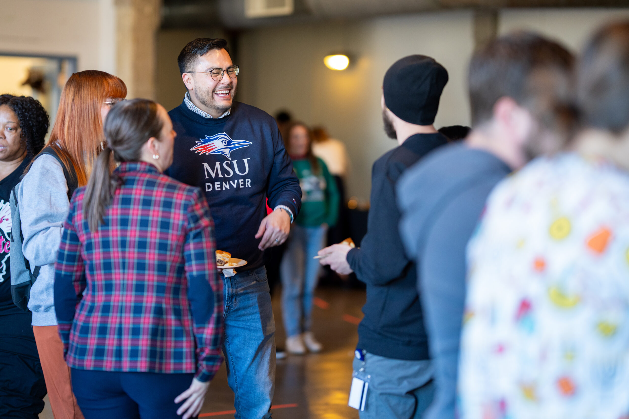 A person in an MSU Denver sweatshirt smiles and chats with others in a festive gathering.