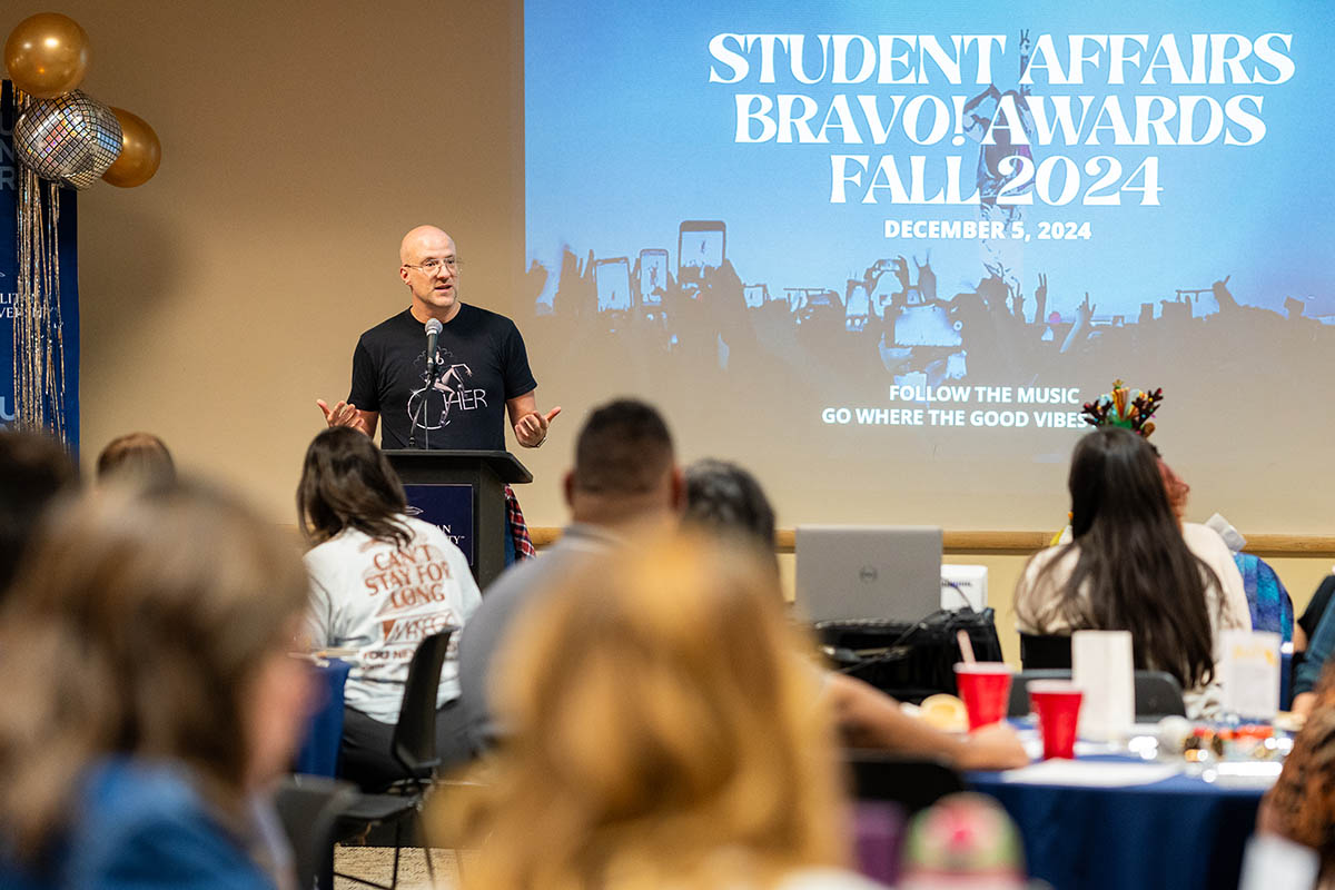 A speaker at a podium addresses a seated audience in a decorated room. The screen behind displays the text "Student Affairs BRAVO Awards Fall 2024."