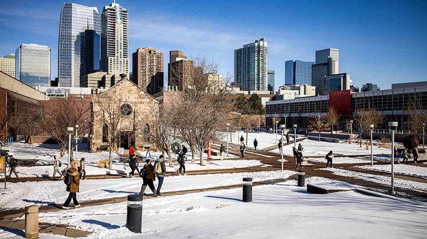 Students walk along snowy pathways on a college campus with a stone chapel and tall skyscrapers in the background on a sunny winter day.