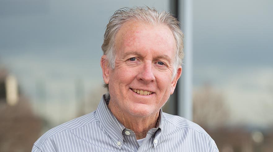 A middle-aged man with light skin and gray hair wearing a striped button-up shirt, smiling warmly at the camera. The background shows an outdoor setting with blurred trees and a cloudy sky.