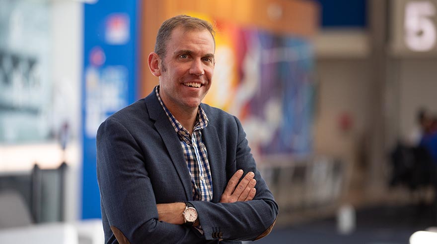 A smiling man in a blue blazer and plaid shirt stands with arms crossed in a brightly lit space, featuring blurred colorful artwork and modern decor in the background.