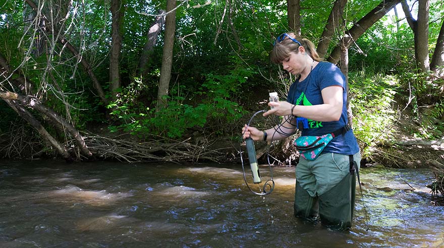 A person in waders stands in a shallow stream surrounded by trees, using scientific equipment to collect water samples.