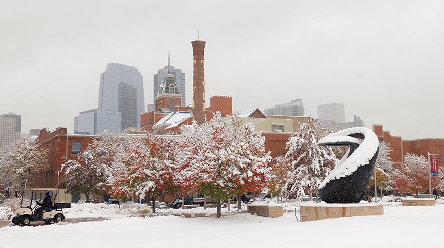 A snow-covered campus with colorful autumn trees contrasts with red brick buildings and the Denver skyline in the background. A distinctive white and black sculpture stands in the foreground.