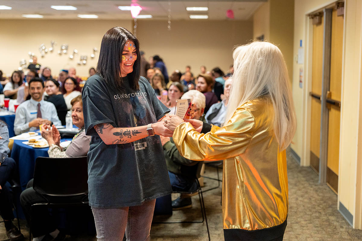 A smiling woman with face paint receives an award from another person wearing a gold jacket, as the seated audience watches in the background.