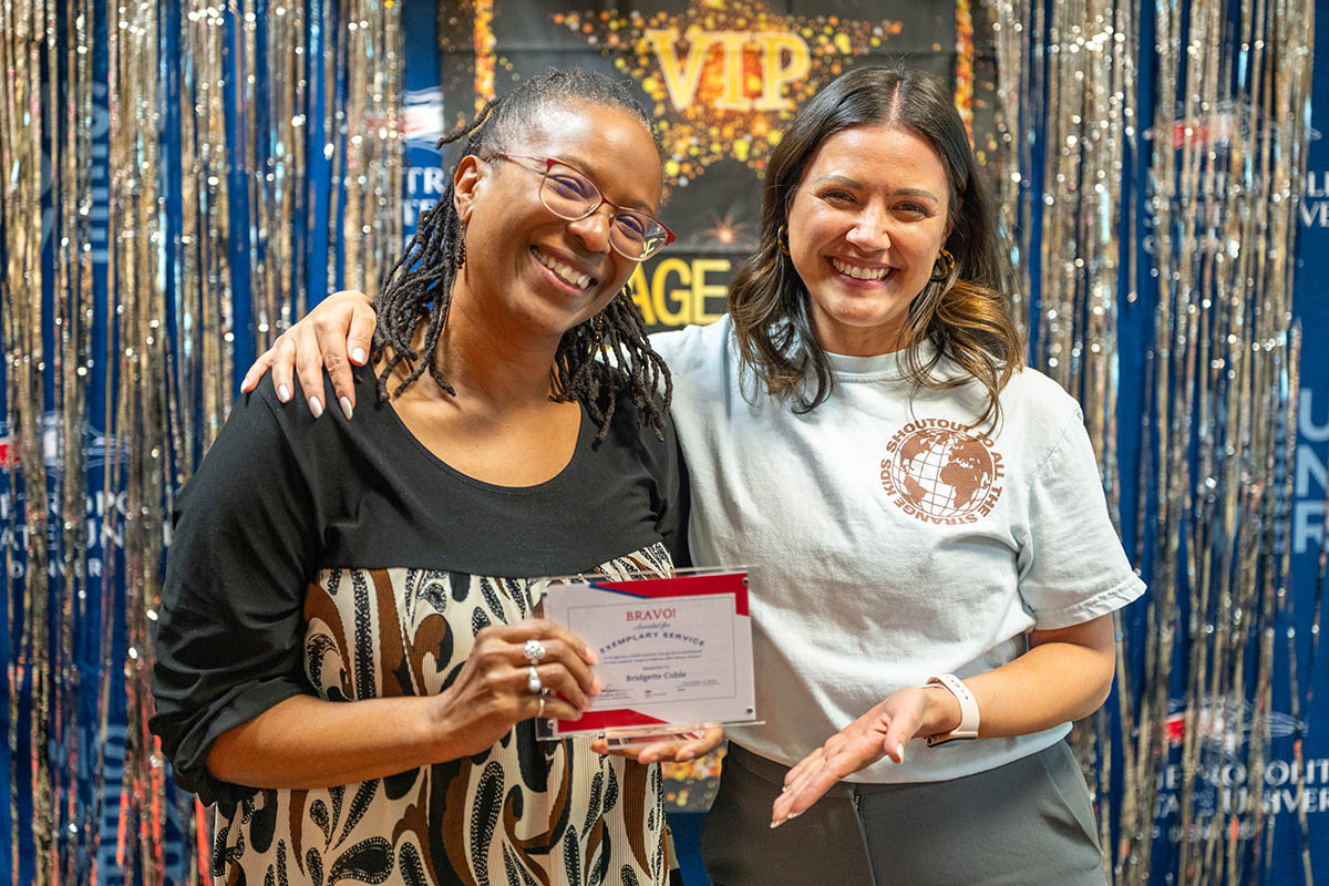 Two women stand together, smiling, as one holds a certificate. A festive backdrop with streamers and "VIP" signage is behind them.