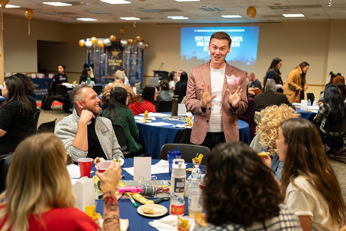 A man in a pink blazer speaks animatedly with attendees seated at round tables during the awards ceremony.