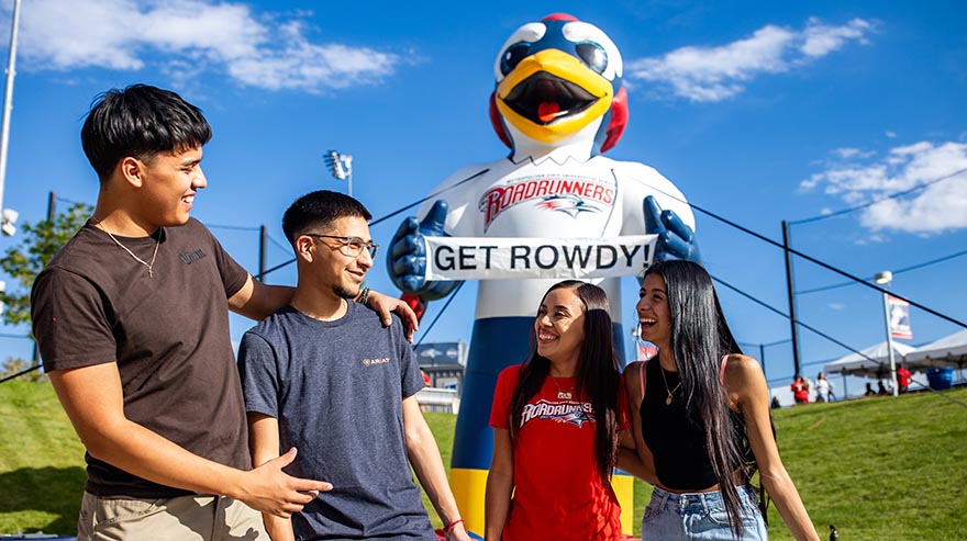 Four students laughing together in front of a large inflatable roadrunner mascot holding a "GET ROWDY!" sign at an outdoor campus event.