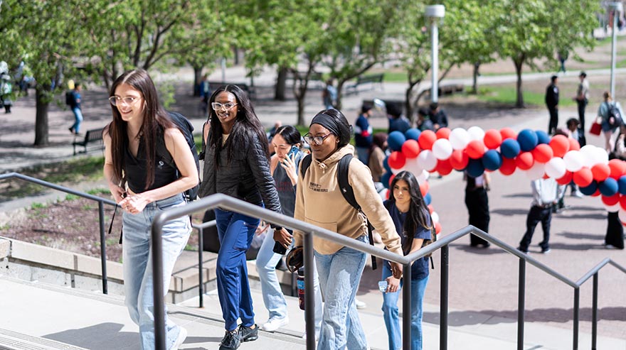 Students walk up stairs on campus near a red, white, and blue balloon arch during a sunny outdoor event.