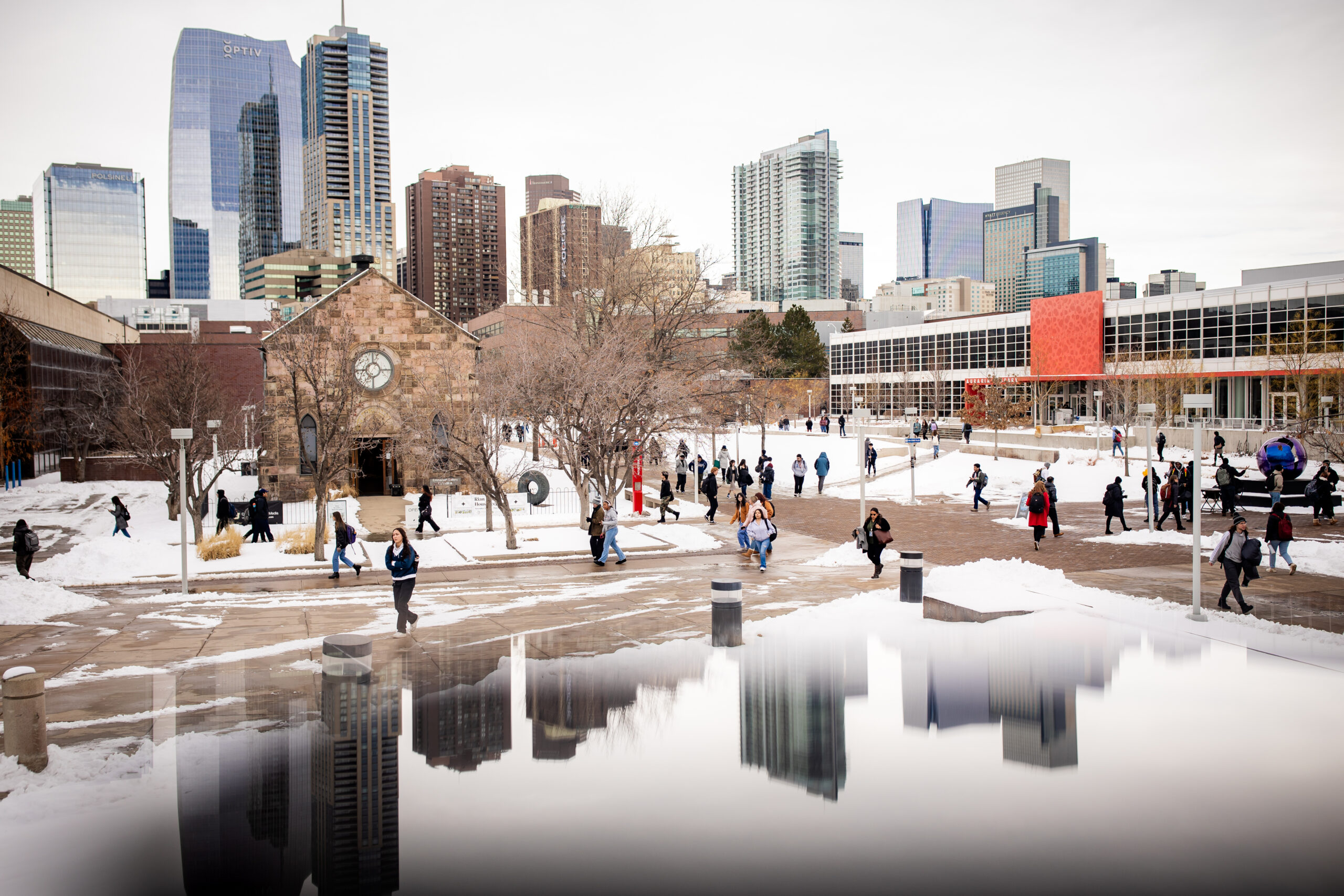 Students walk through a snowy campus with a historic chapel and Denver's skyline in the background, reflected in a foreground surface.