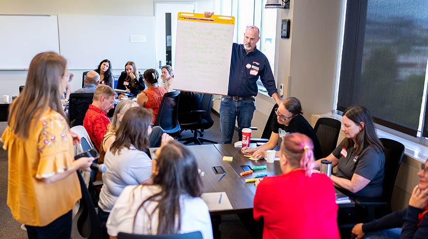 A collaborative workshop in a classroom, with a man holding a large notepad while participants engage in discussion around tables.