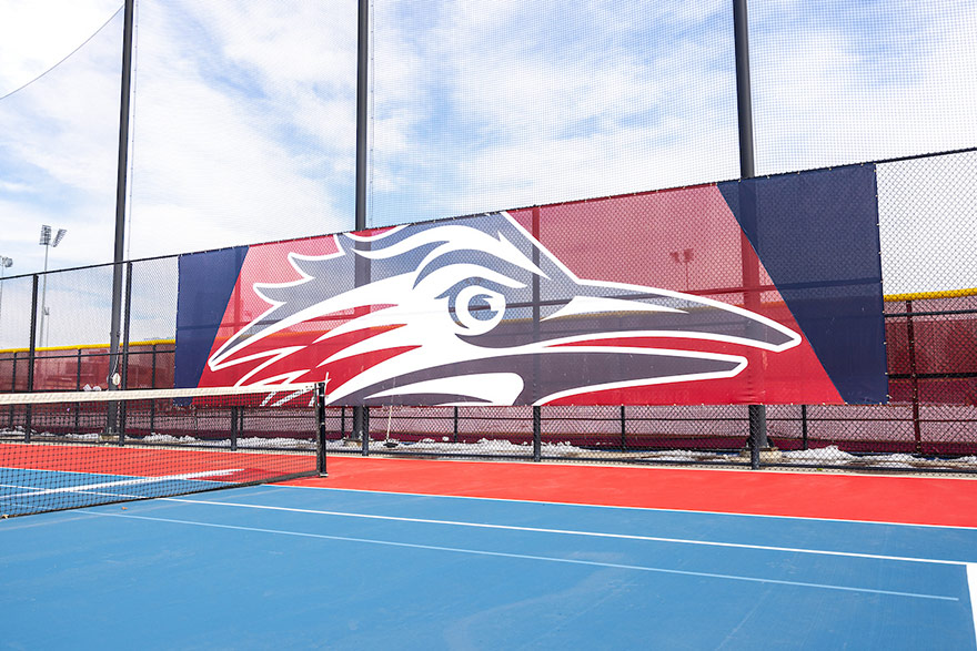 Photo of tennis court. A large banner with the MSU Denver Roadrunners logo appears on a fence behind the court.