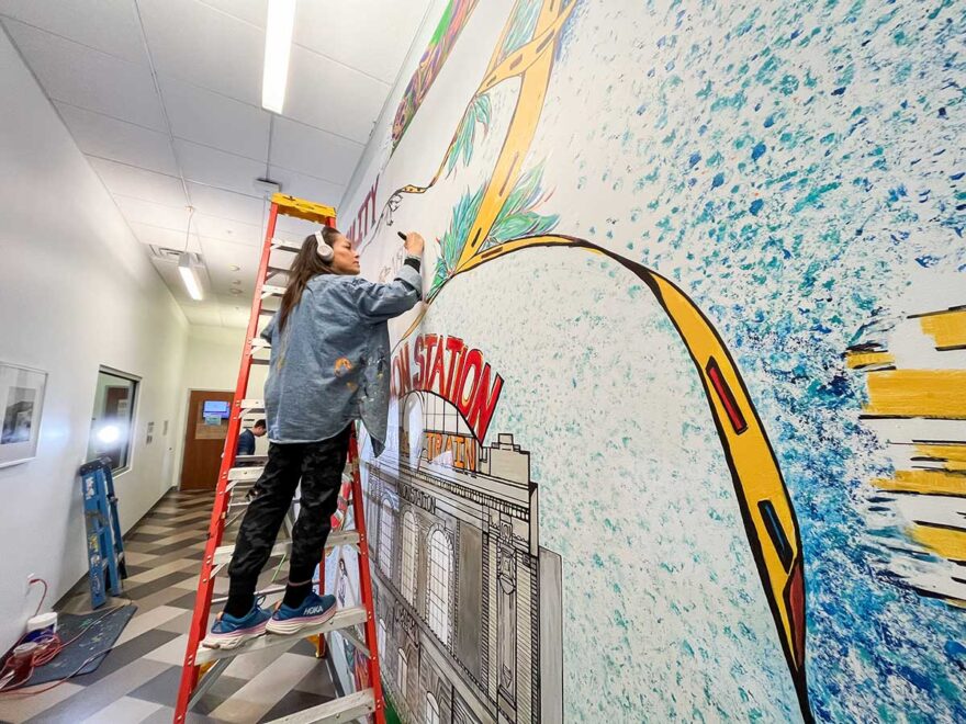 Artist Lisa Lopez Huffman stands on a step ladder and paints part of a large indoor mural that includes an image of Denver's Union Station.