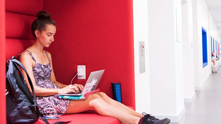 A person works on a laptop in a red cushioned study nook in a modern, well-lit hallway with similar study spaces.