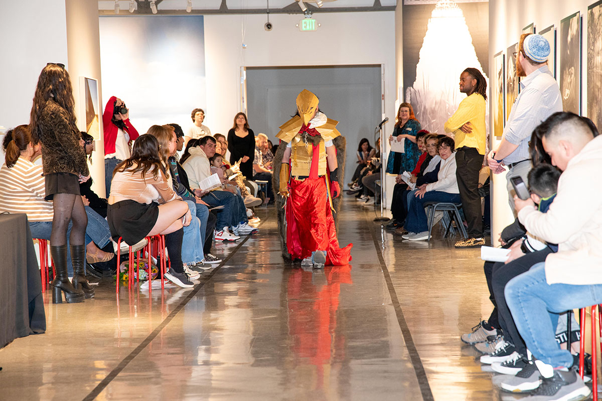 A model in a golden warrior costume walks down the Art + Action fashion show runway, with an audience watching.