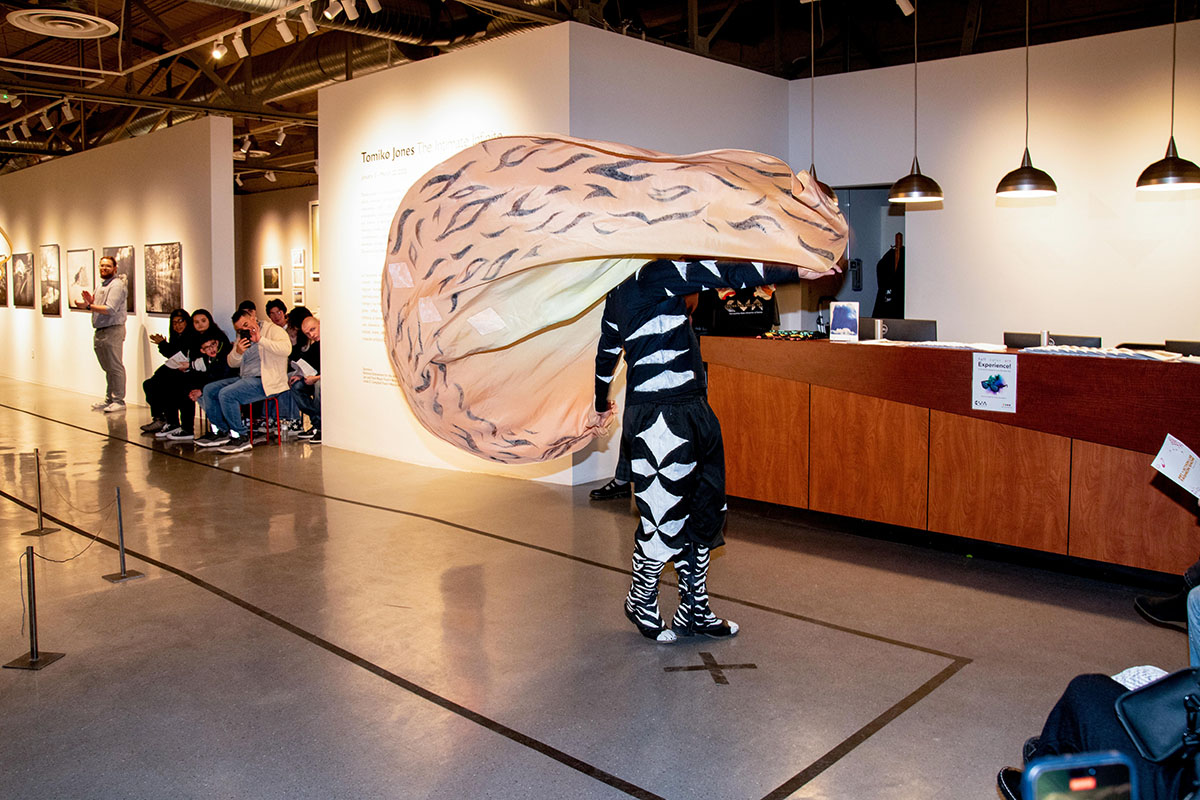 A performer showcases a dramatic fashion piece with oversized, flowing fabric that mimics organic textures. The outfit is black and white with bold zebra-like patterns, and the large fabric element billows as they move through the gallery.