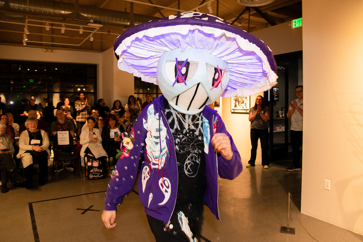 A model in a glowing mushroom headpiece and a colorful outfit walks the Art + Action fashion show runway.