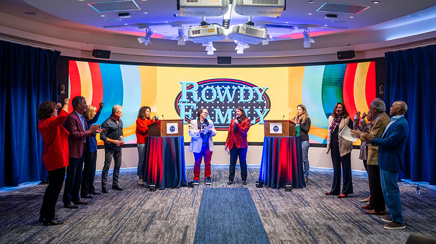 "Rowdy Family" game event with two podiums and a colorful screen. Two women stand between the podiums while participants on both sides clap and cheer.