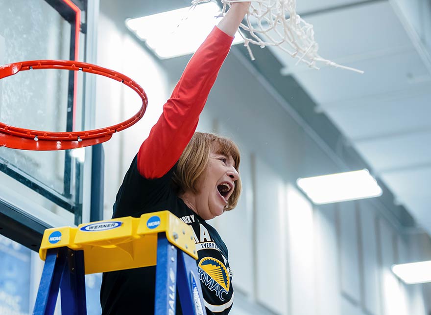 Basketball coach Tanya Haave stands on a ladder and raises a net, cut down from the hoop, in the air as she celebrates a tournament win.