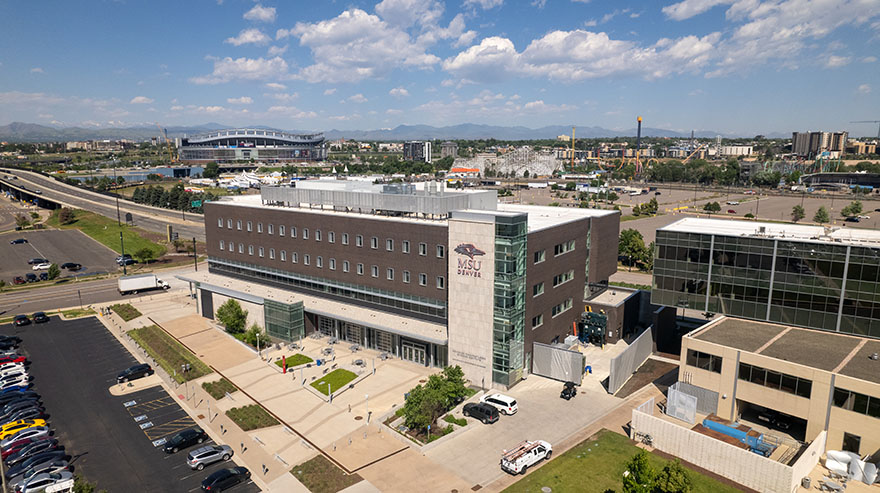 An aerial view of the MSU Denver Aerospace and Engineering Sciences (AES) Building on a sunny day. The modern structure features a mix of dark panels and glass, with the MSU Denver logo prominently displayed on a light-colored tower. The surrounding area includes parking lots, landscaped walkways, nearby buildings, and a distant view of downtown Denver, with the mountains and stadium visible on the horizon.