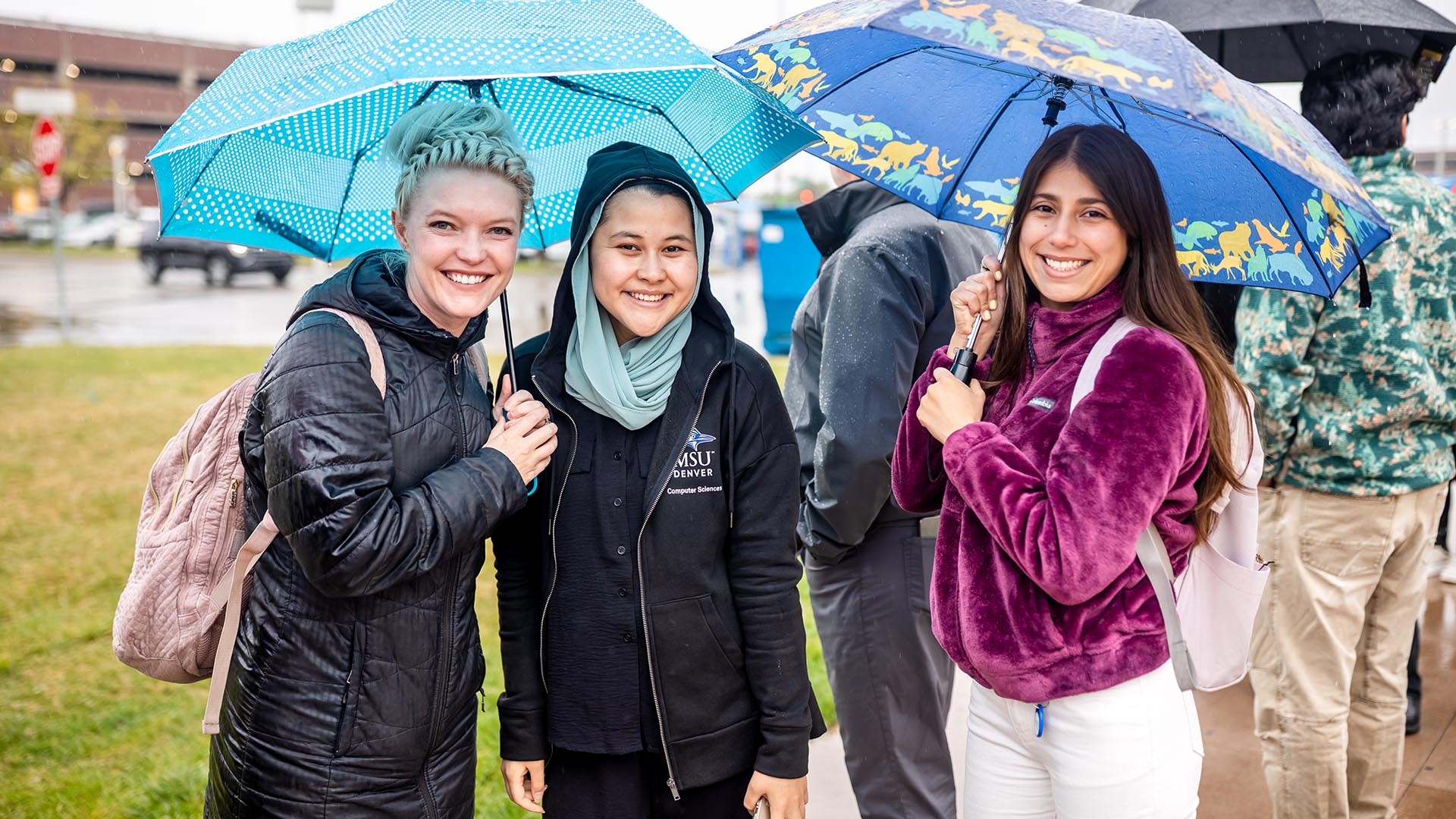 Three individuals stand close together under umbrellas on a rainy day, smiling while holding umbrellas decorated with polka dots and animal prints. An MSU Denver Computer Sciences jacket is visible among the group.