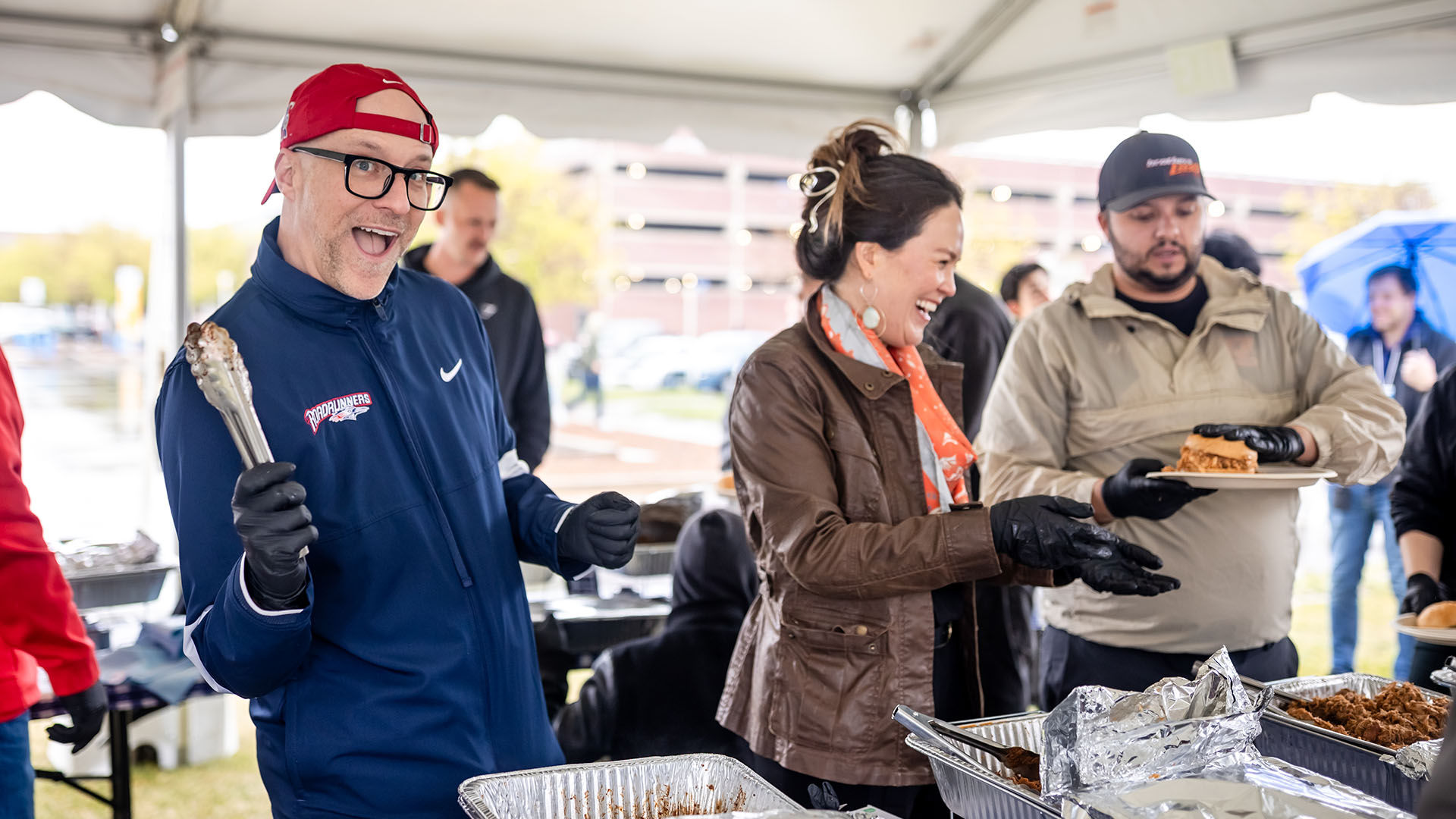 Participants serve barbecue food under a tent, smiling and laughing. One person enthusiastically holds tongs over a tray of food while others help serve or receive plates.