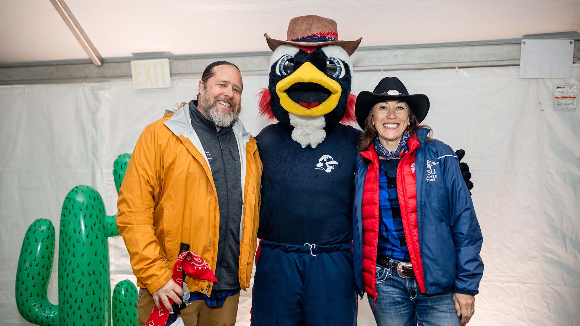 Dr. Matt Makley, the MSU Denver mascot Rowdy wearing a cowboy hat, and Dr. Janine Davidson pose together inside the event tent during the Howdy Rowdy BBQ. They stand in front of a white backdrop with green inflatable cacti nearby.