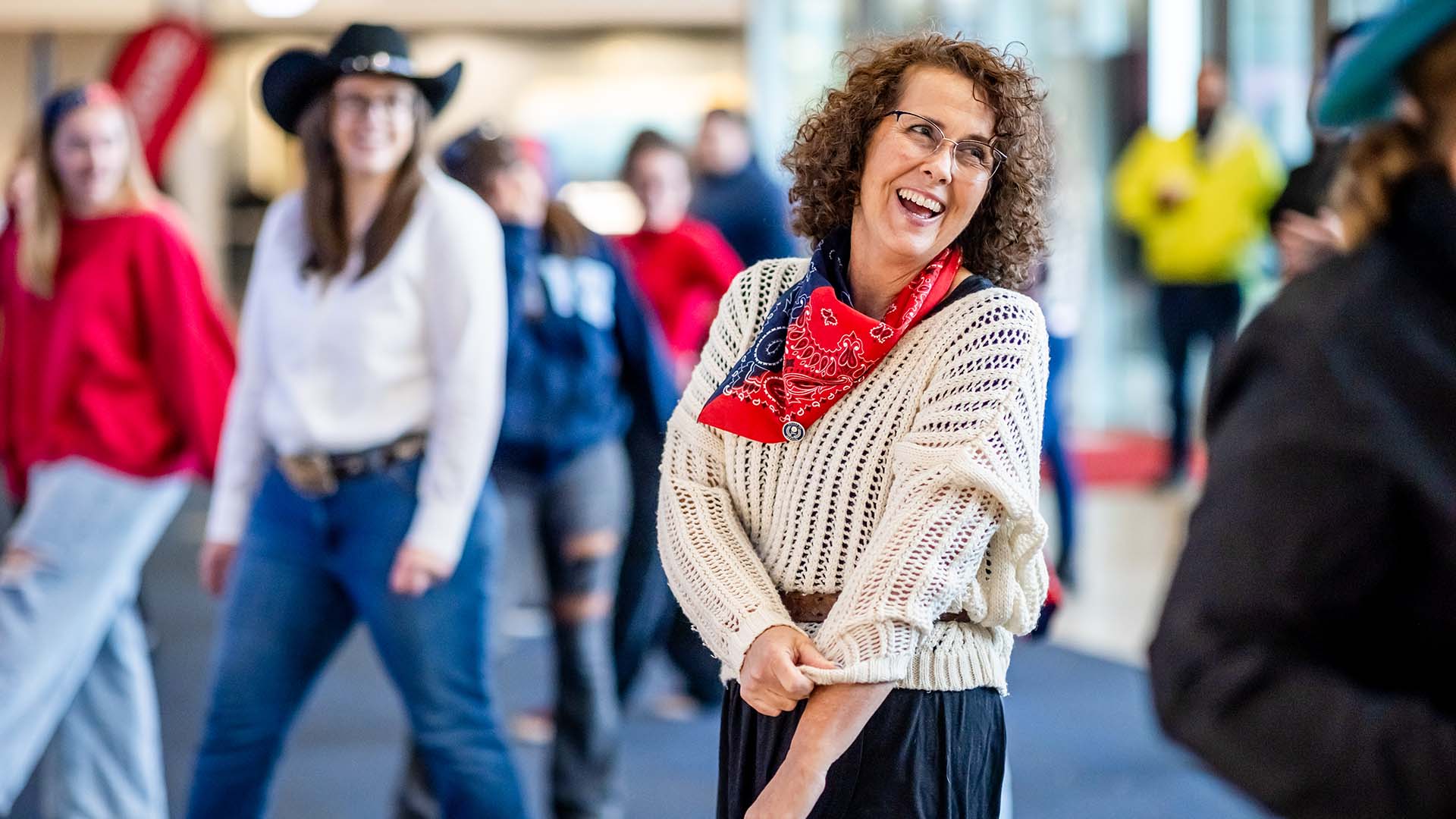A participant with curly hair, a white knit sweater, and a red bandana smiles while line dancing in a large indoor space. Others dressed in western attire join in the background.