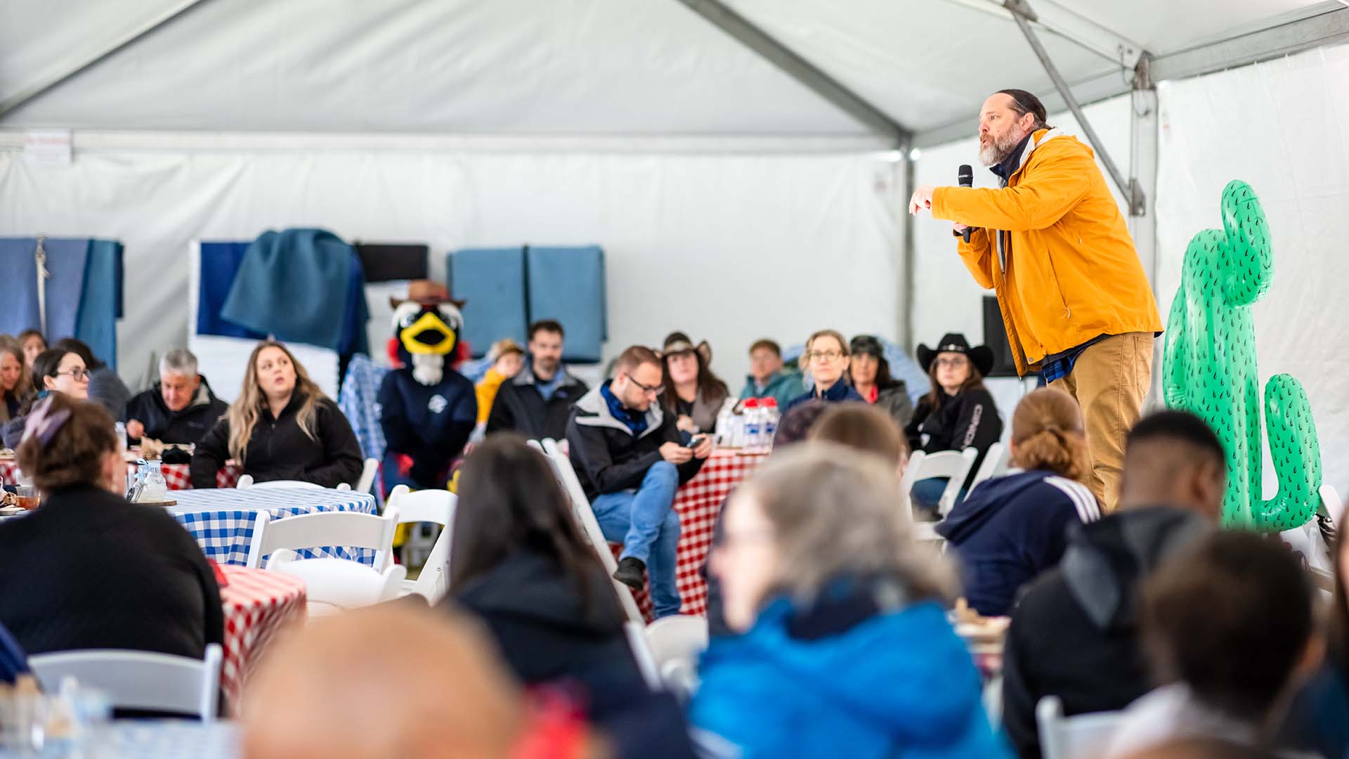 Matt Makley, wearing a yellow jacket, speaks into a microphone while addressing a seated audience inside a large event tent during the Howdy Rowdy BBQ. Inflatable green cacti decorate the stage, and the MSU Denver mascot, Rowdy, is visible among attendees in the background.