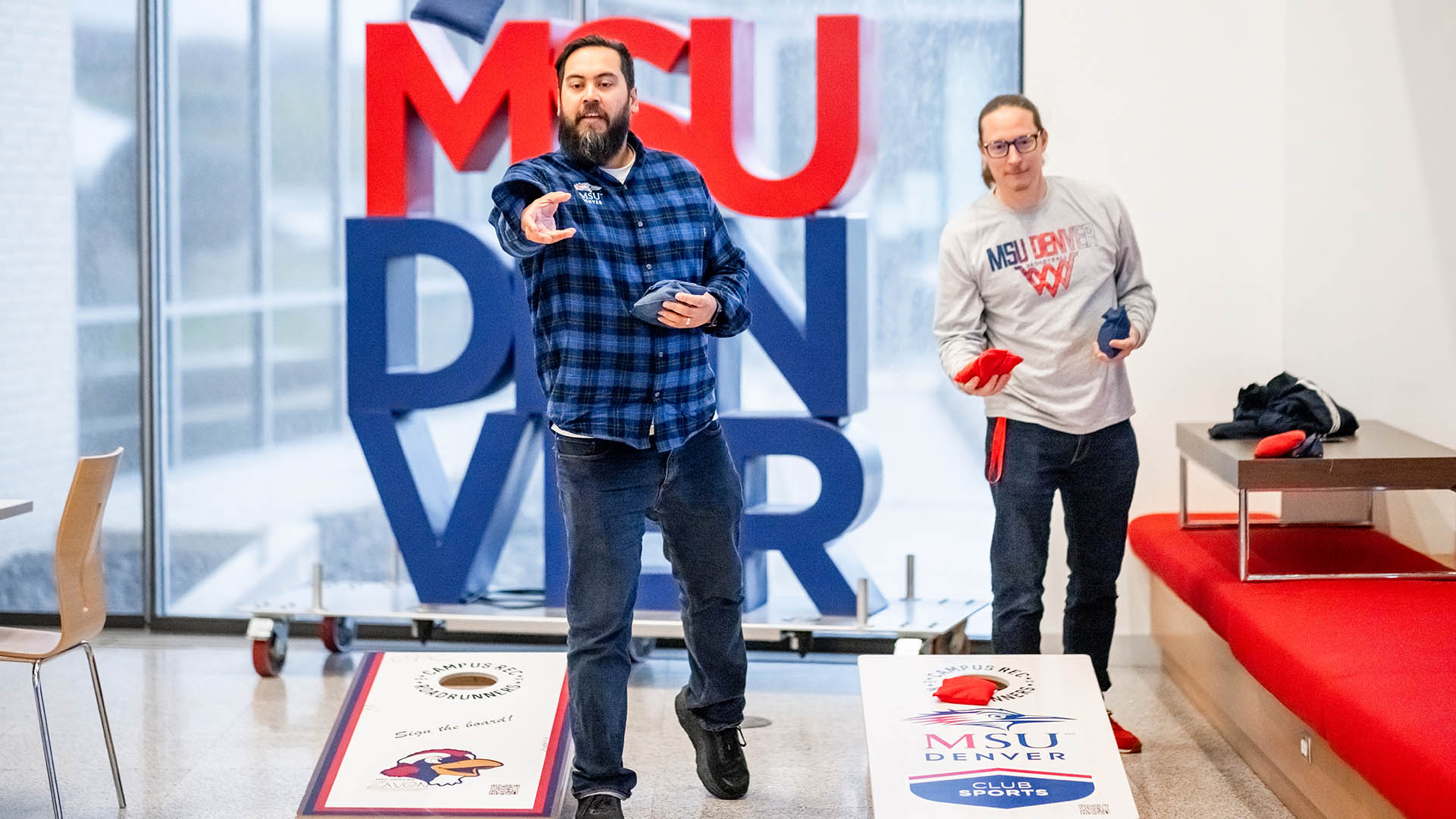 Two individuals play cornhole inside a bright indoor space, with large red and blue letters spelling “MSU Denver” in the background. Each person holds bean bags while aiming at boards branded with the university’s Roadrunners logo.