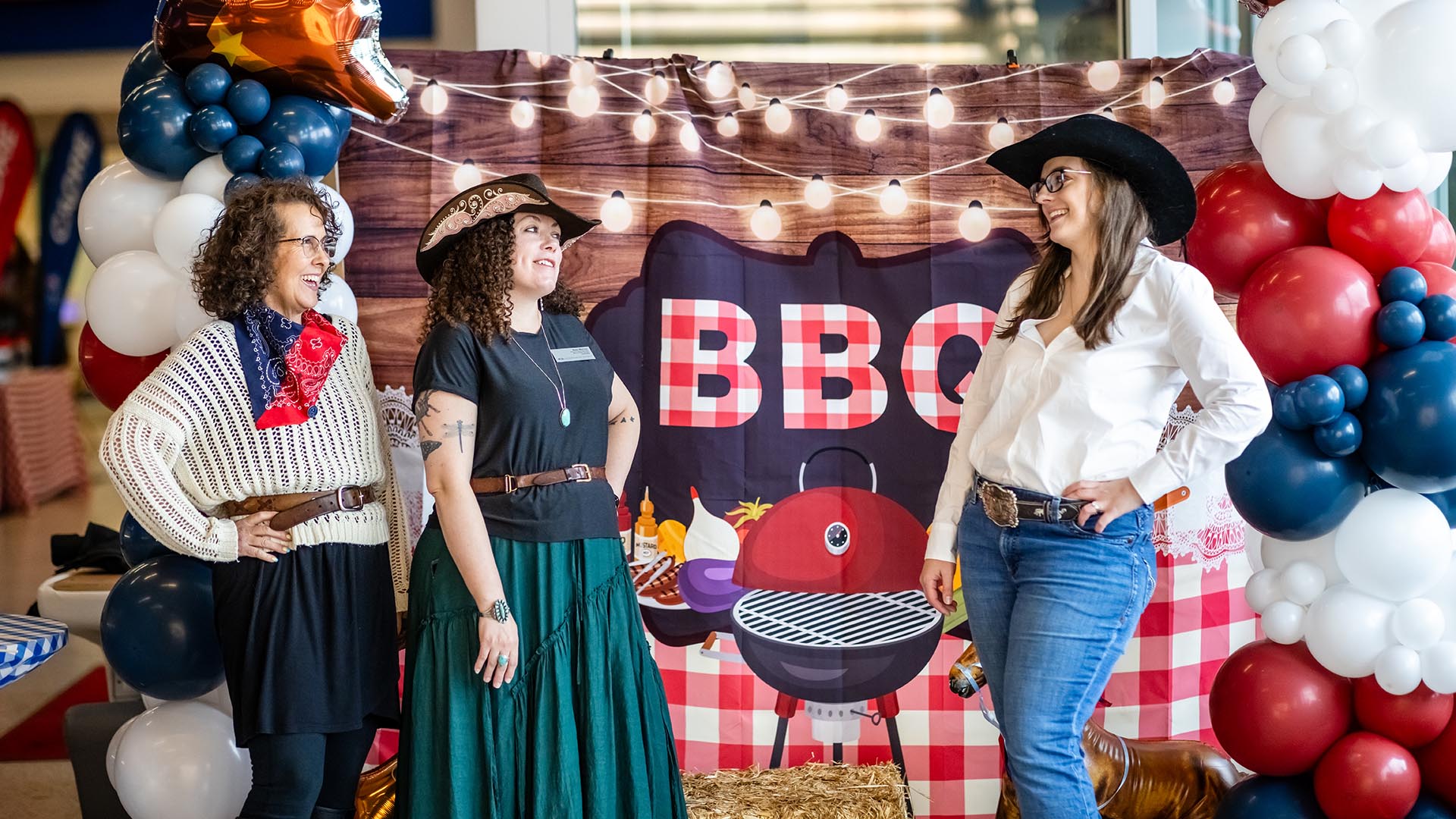 Three participants, dressed in western-themed clothing including cowboy hats and bandanas, stand smiling in front of a festive “BBQ” photo backdrop. The display includes balloons in red, white, and blue, along with hay bales and string lights.