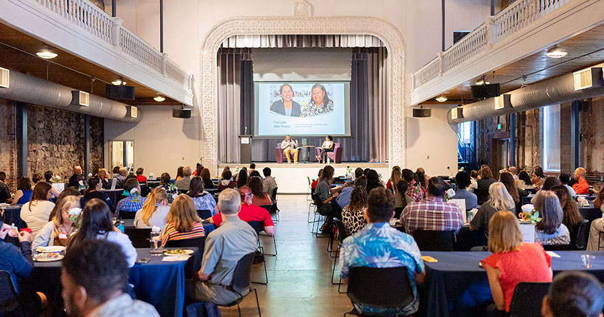 A large group of people sit at tables facing a stage with two speakers during a community event.