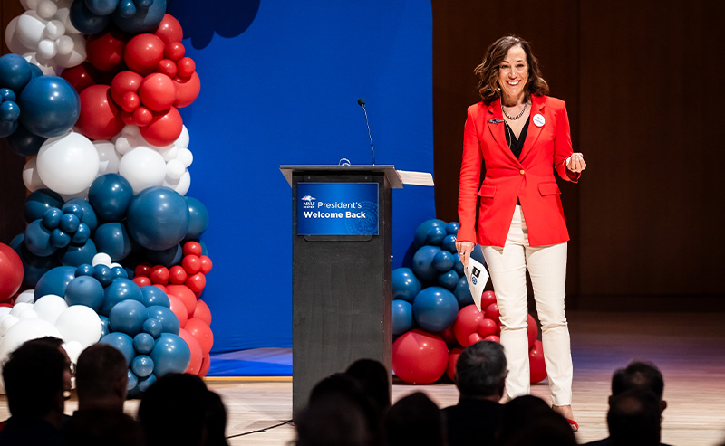 MSU Denver President Janine Davidson speaks on stage during the “President’s Welcome Back” event, wearing a red blazer and smiling toward the audience, with a podium and red, white, and blue balloon decorations in the background.