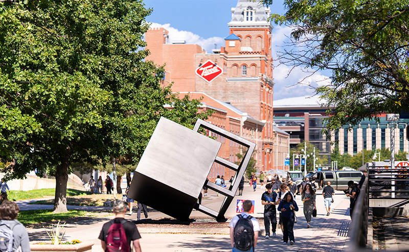 Students walking near the tilted cube sculpture with the Tivoli Student Union in the background on the Auraria Campus.