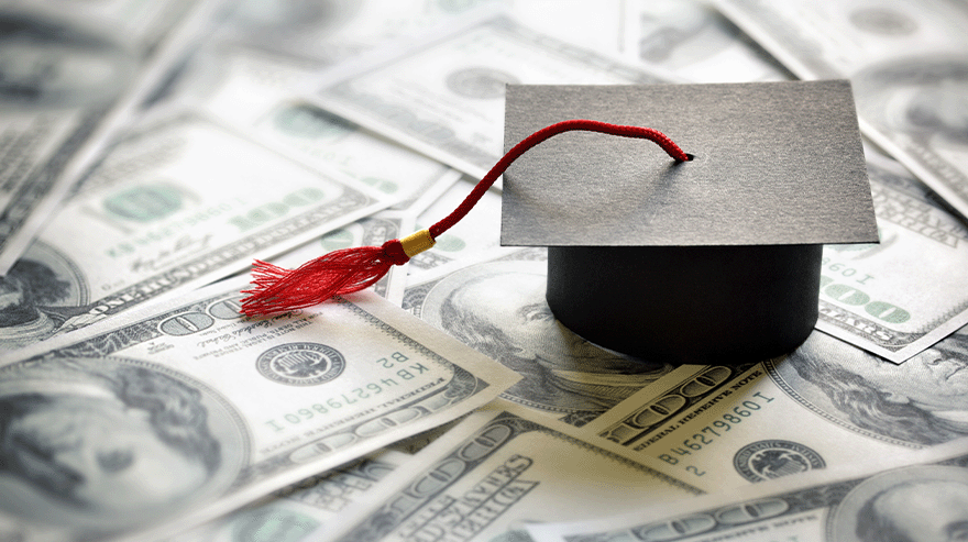 Miniature graduation cap with red tassel placed on top of scattered U.S. hundred-dollar bills, symbolizing student loans and education costs