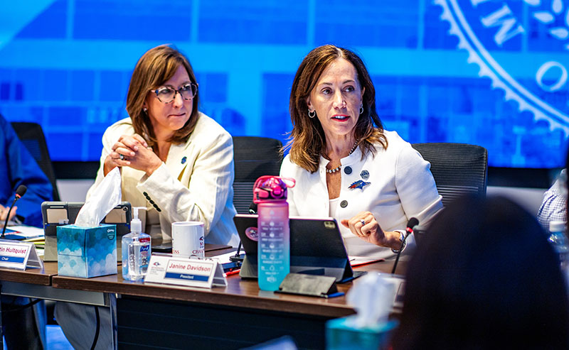 Two people in white blazers seated at a boardroom table, one speaking while the other listens.
